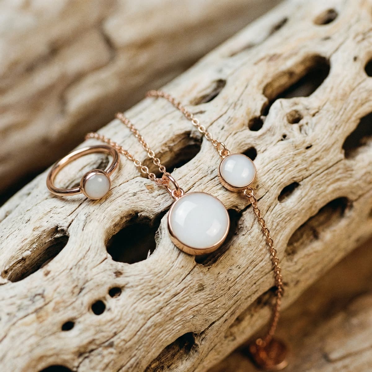 Gioielli ricordo per il fine allattamento: collana, anello e bracciale color oro rosa con cabochon bianco latte, fotografati su legno naturale.