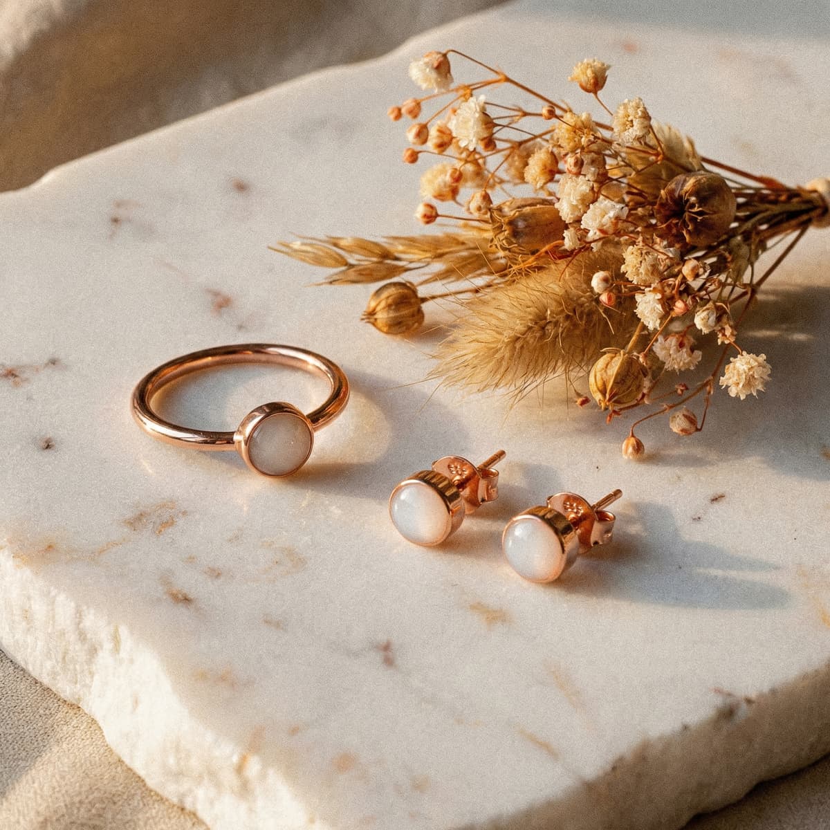 Set regalo elegante con anello e orecchini a lobo in oro rosa con pietra bianca lattiginosa su base in marmo accanto a fiori secchi, miglior regalo neo mamma per un ricordo prezioso.
