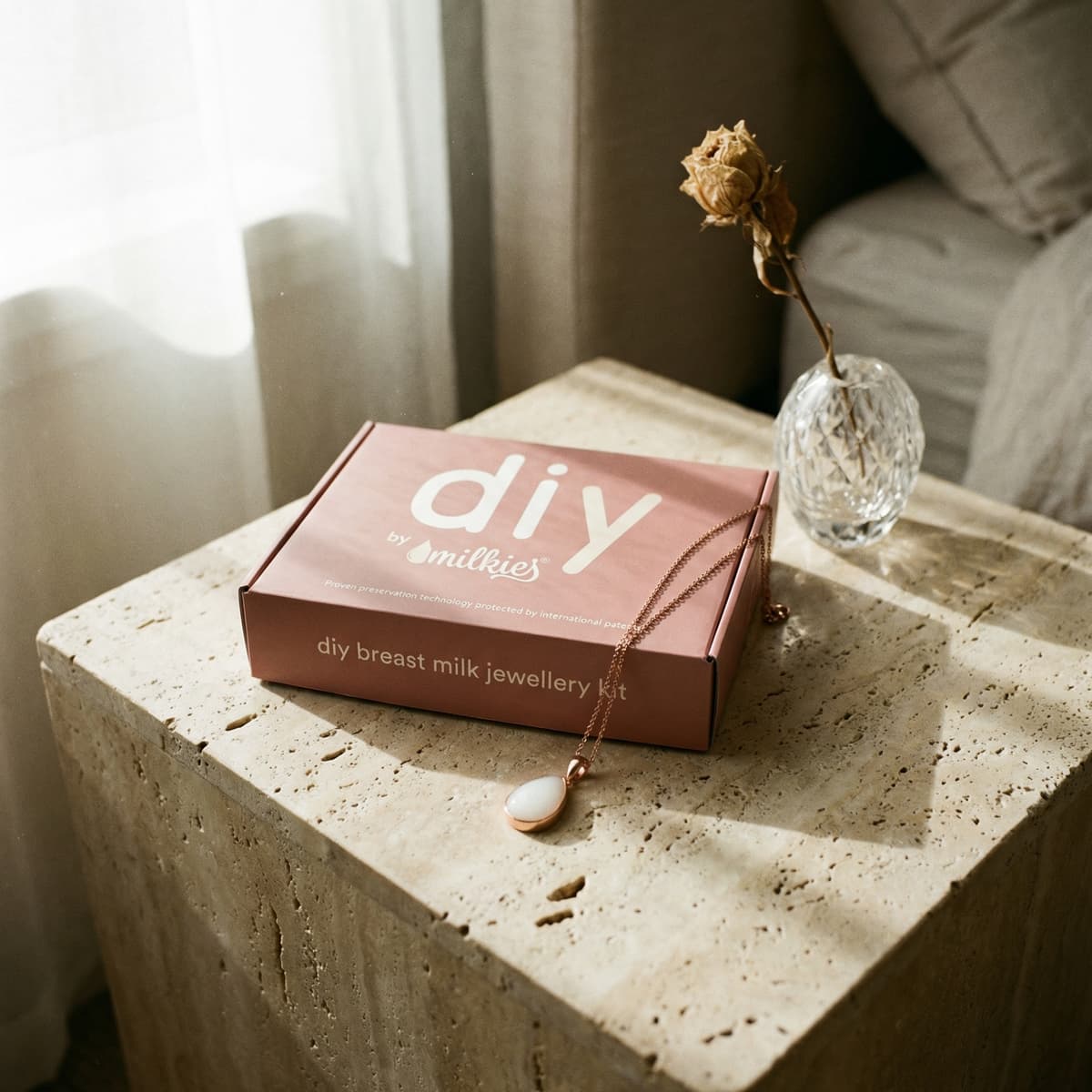 Scatola rosa del kit DIY by MILKIES su tavolino in pietra con collana pendente e vaso con fiore secco, ideale come regalo nascita mamma per creare gioielli con latte materno.