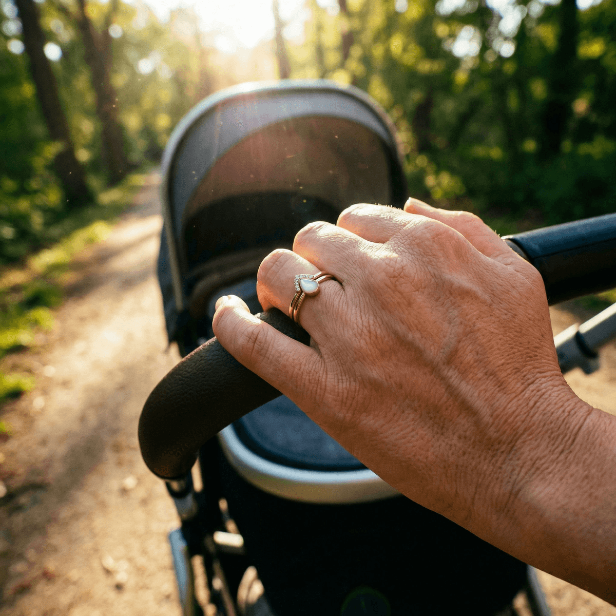 Mano di una mamma che spinge un passeggino in un parco al tramonto, indossando eleganti anelli latte materno in oro rosa con pietra bianca a goccia e piccoli cristalli.