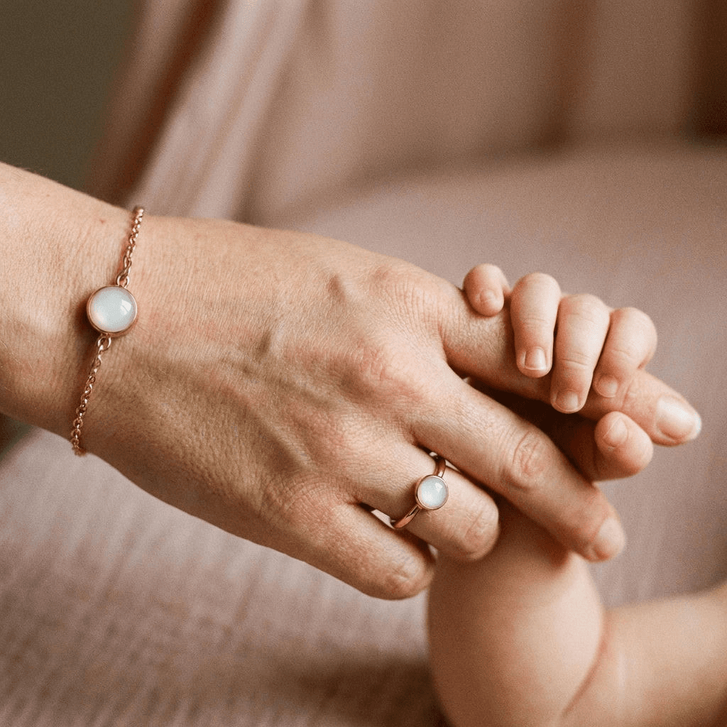 Mano di una mamma che tiene la mano del bambino, con un bracciale con latte materno fai da te e un anello coordinato in oro rosa con pietra lattiginosa.