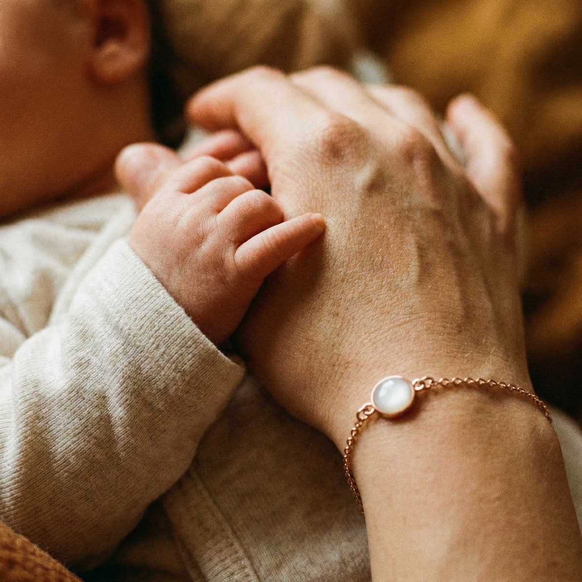 Mano di mamma che tiene un neonato mentre stringe un delicato bracciale latte materno in catena oro rosa con ciondolo rotondo bianco effetto perla.