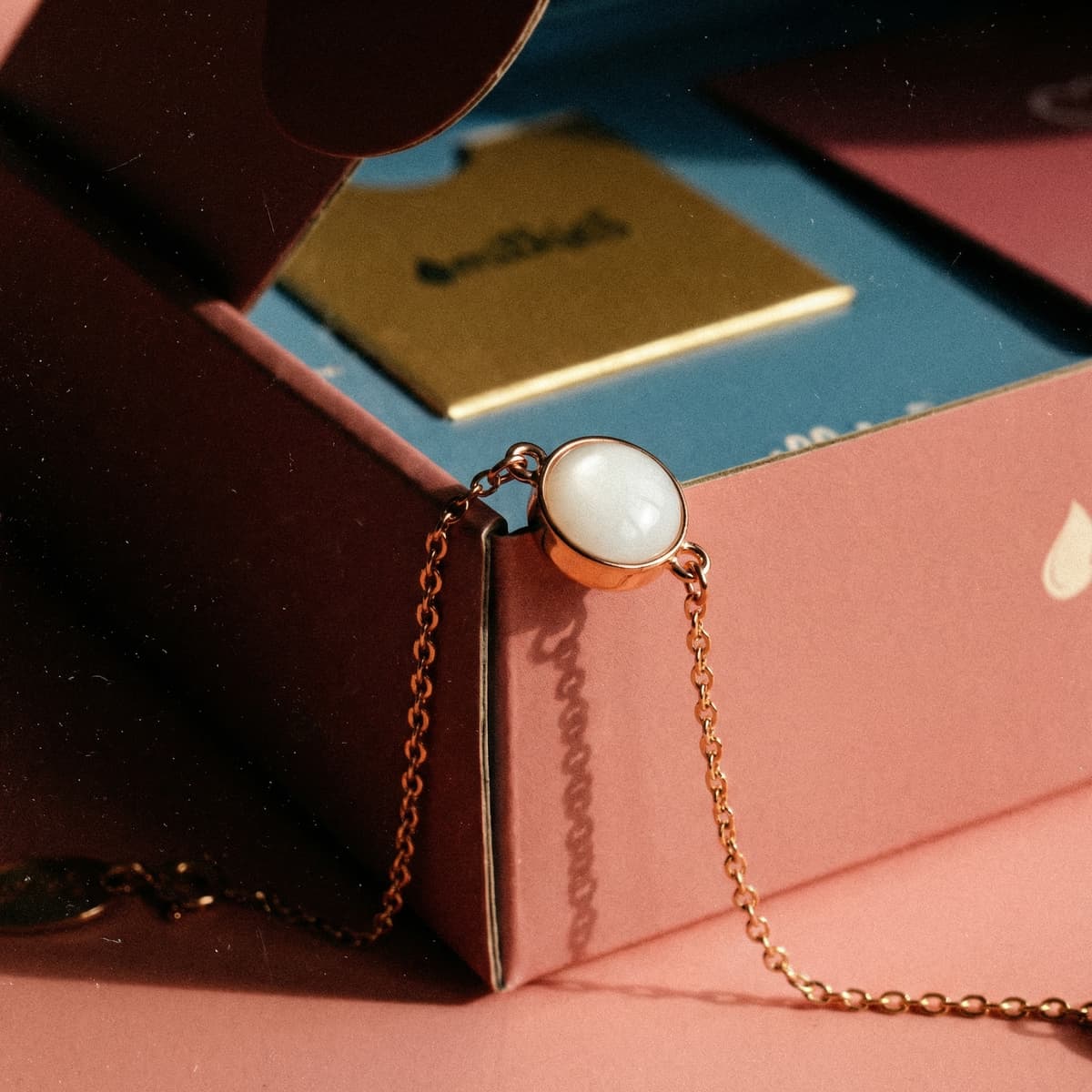 Bracciale in catena color oro rosa con cabochon bianco opalescente, un bracciale latte materno fai da te, appoggiato su una scatola rosa con il packaging del kit sullo sfondo.