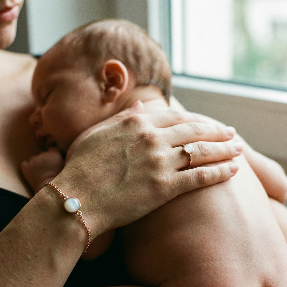 Mamma che coccola un neonato vicino a una finestra, indossando un bracciale latte materno fai da te e un anello con pietra bianca: un ricordo intimo da creare in casa con un kit DIY per chi desidera privacy e controllo sul risultato.