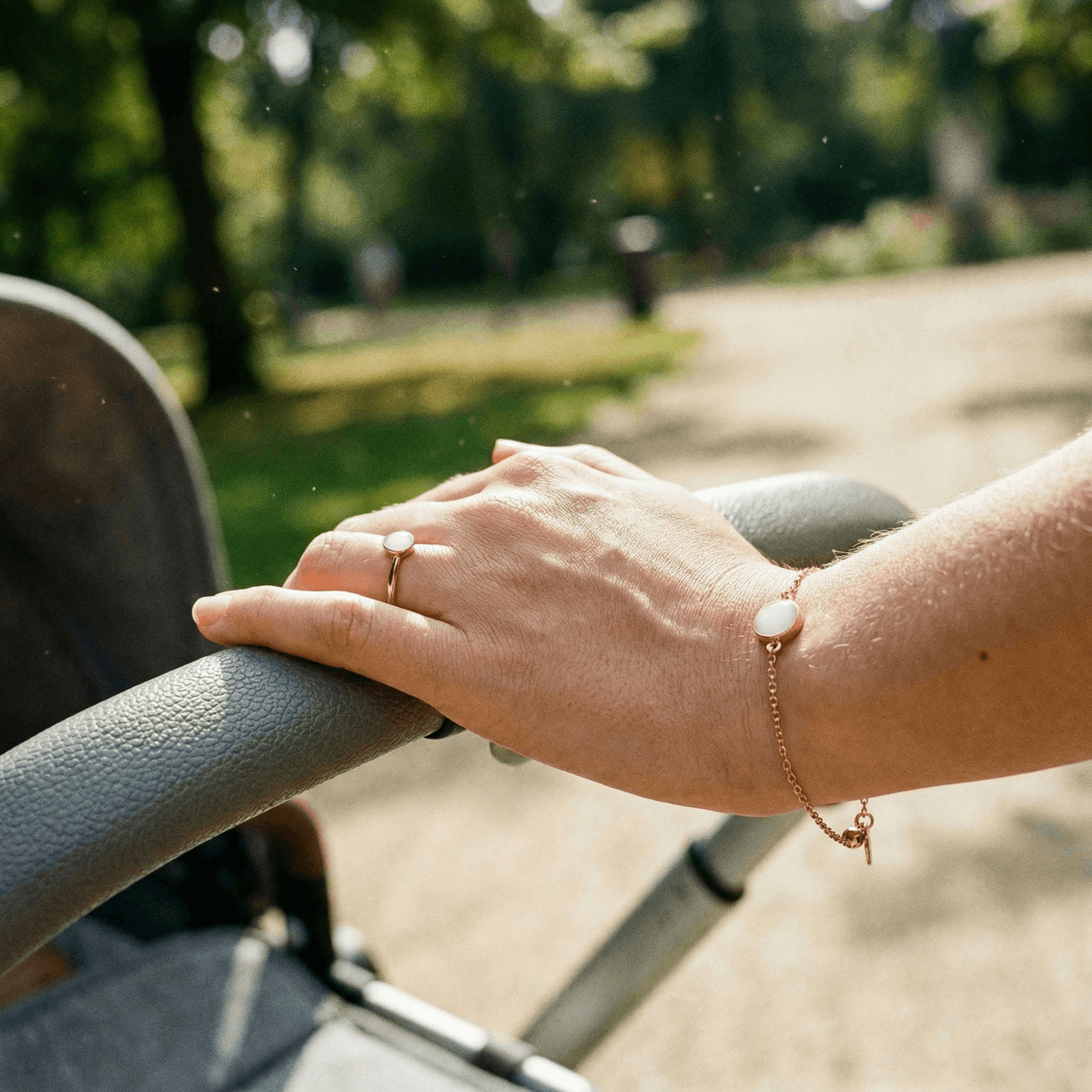 Mano di una mamma che spinge un passeggino al parco indossando un anello e un bracciale in oro rosa con pietra bianca, esempio di gioielli latte materno da indossare ogni giorno
