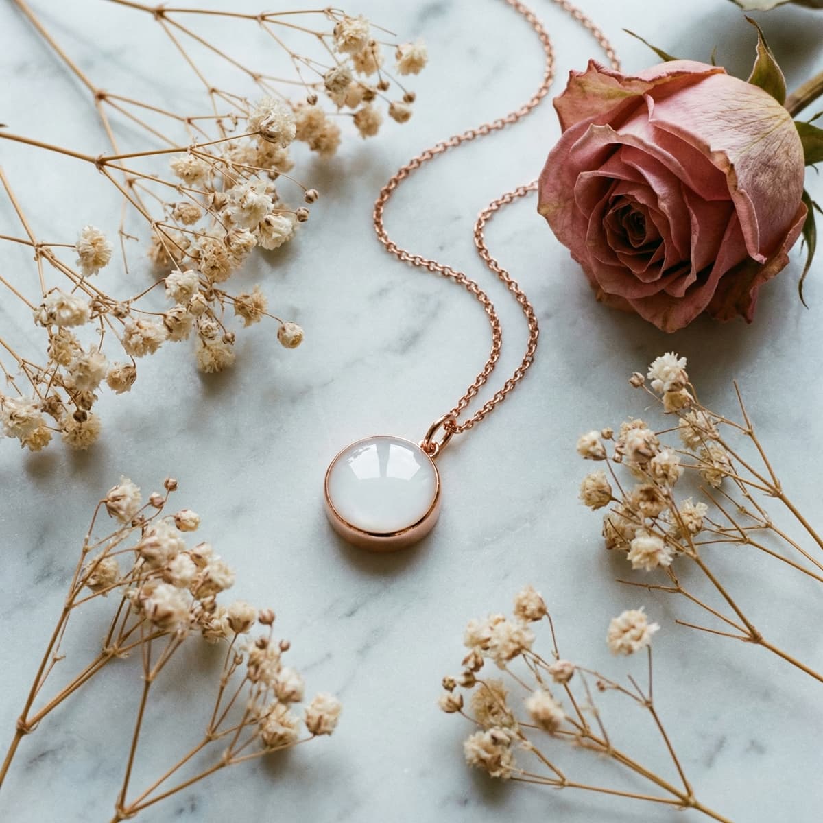 Collana in oro rosa con ciondolo rotondo lattiginoso, ideale come gioiello ricordo latte, appoggiata su marmo con rosellina e fiori secchi