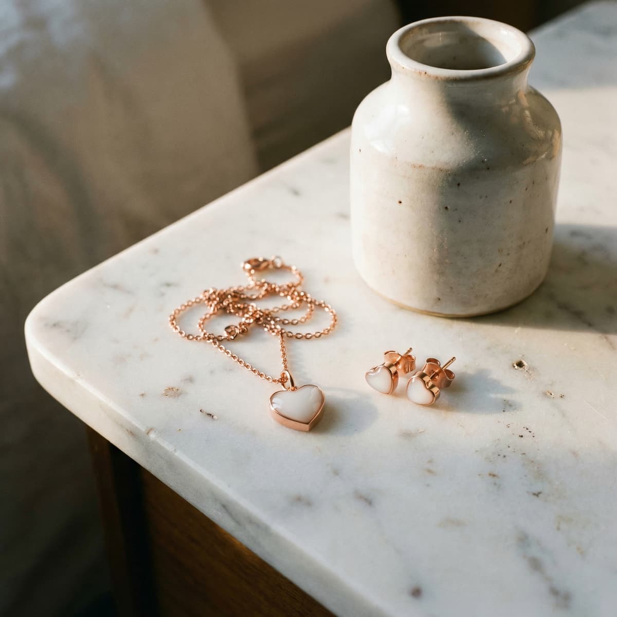 Collana e orecchini a cuore in finitura oro rosa con inserto bianco, esempio di kit fai da te gioielli latte materno, appoggiati su tavolino in marmo accanto a un vaso in ceramica.