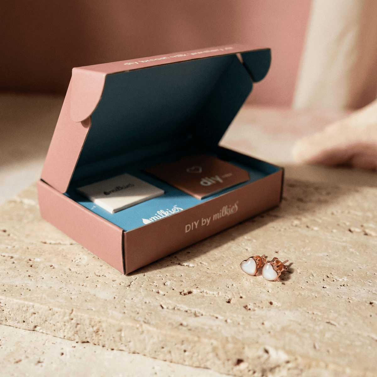 Scatola aperta DIY by MILKIES con materiali e istruzioni e kit orecchini ricordo allattamento, con orecchini a cuore color oro rosa e inserto bianco davanti alla confezione.