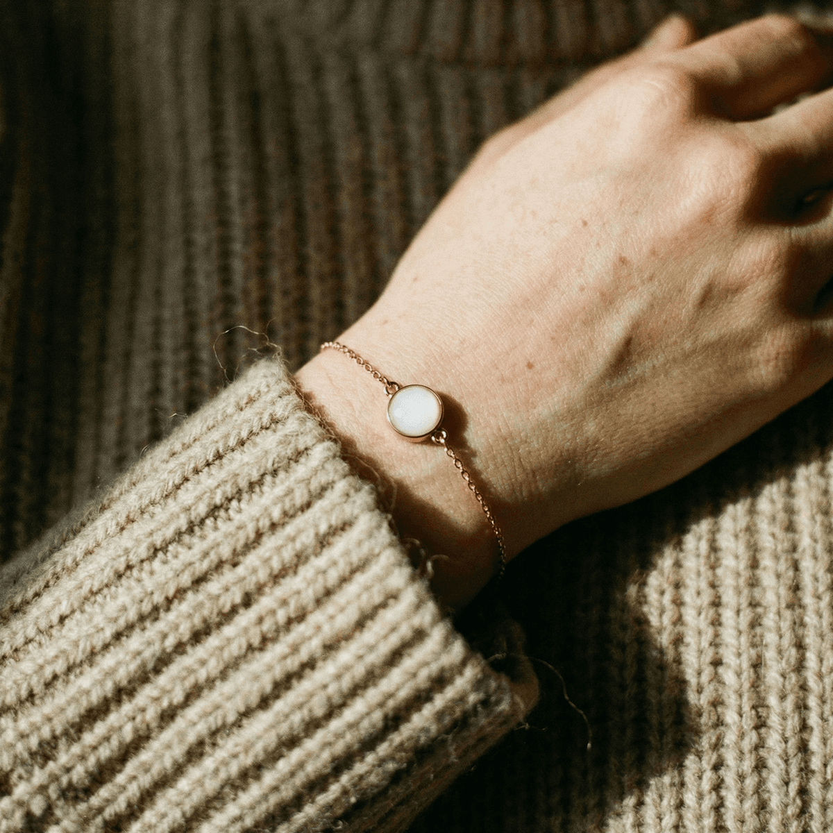 Mano con maglione in lana che indossa un bracciale minimal in catena dorata con cabochon bianco, realizzato con un kit per creare bracciale latte materno come ricordo dell’allattamento