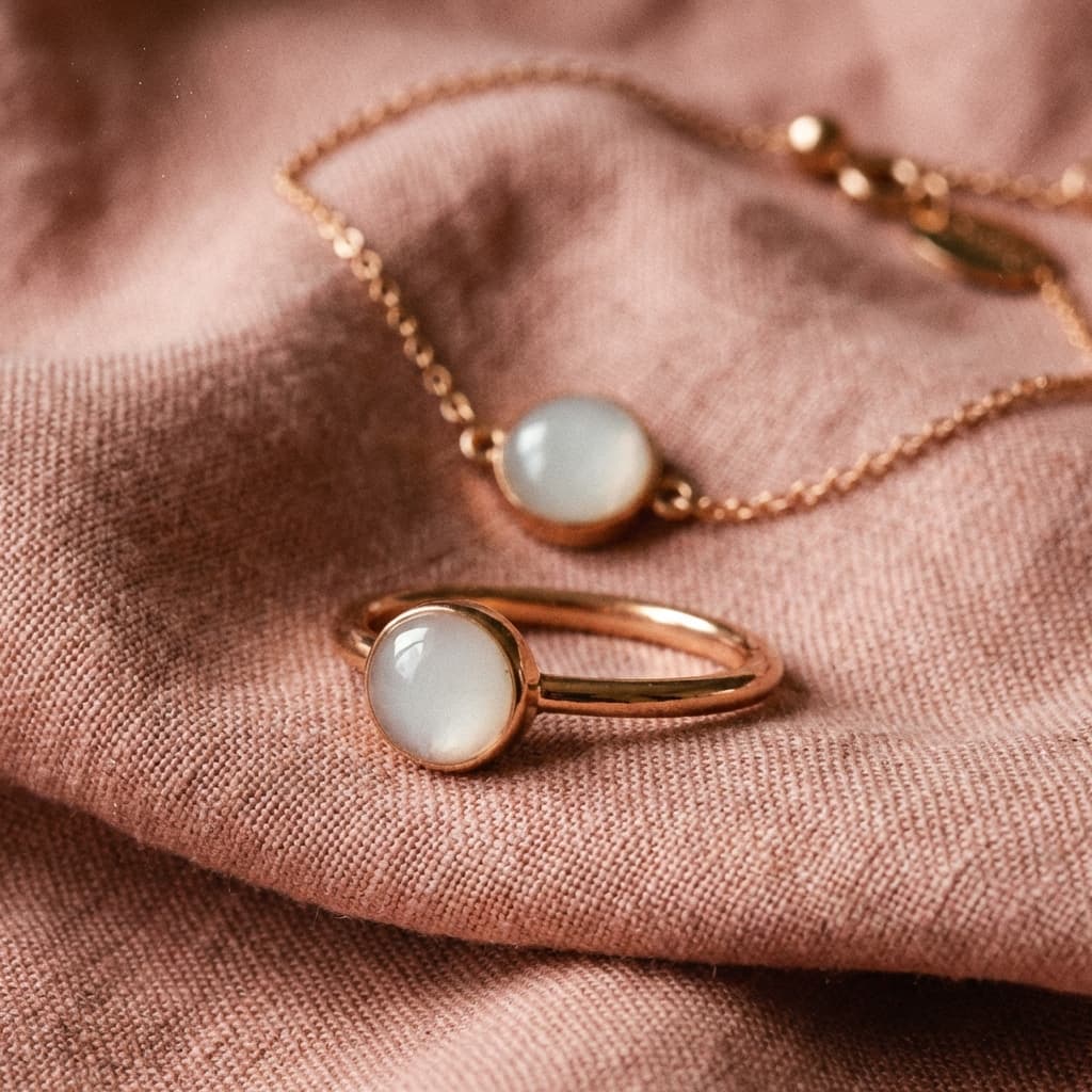 Collana e anello in tonalità oro rosa con pietra bianca lattiginosa, esempio di kit per creare gioielli latte materno, appoggiati su tessuto rosa.