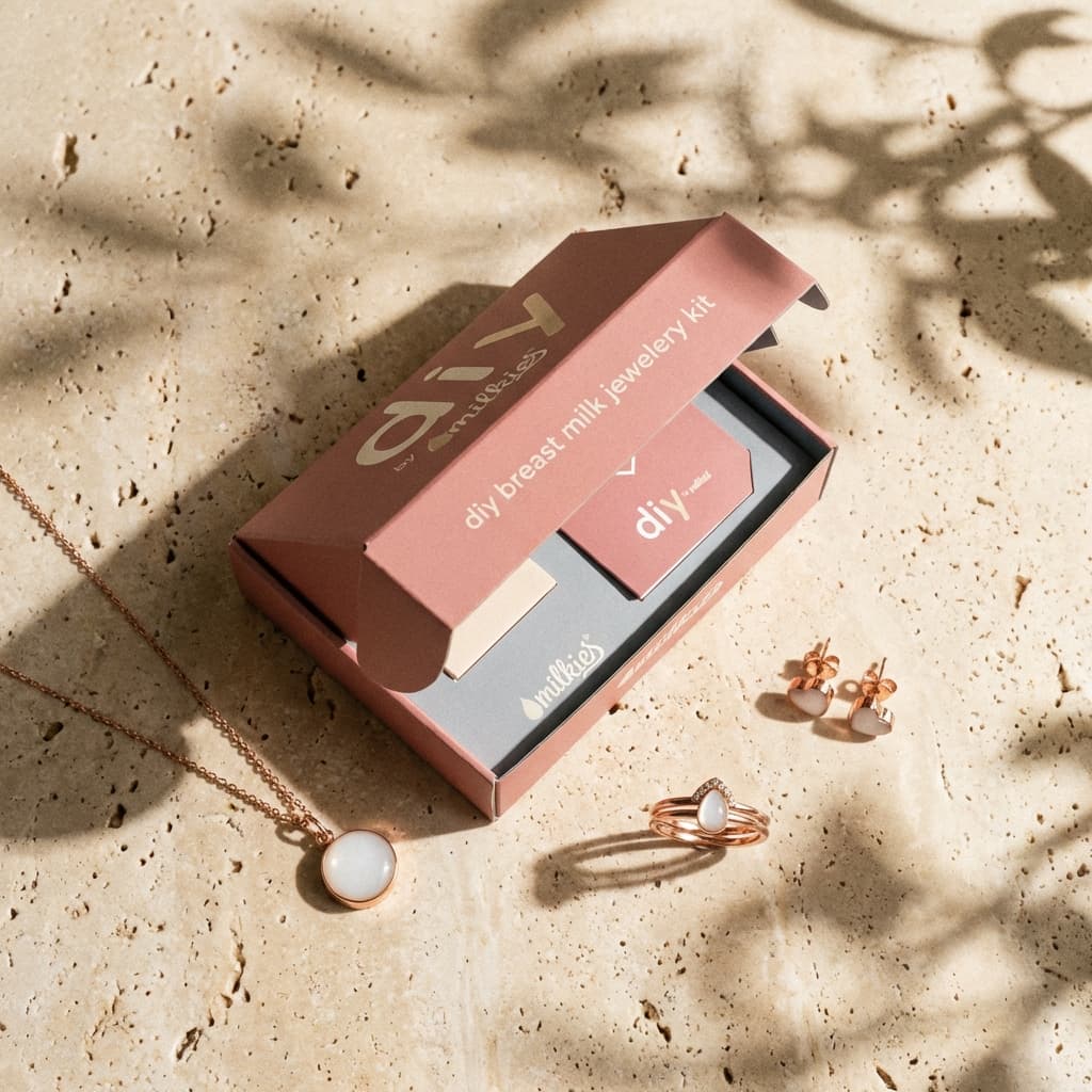 Scatola rosa DIY by MILKIES aperta con kit per creare gioielli latte materno, con collana, anello e orecchini in tonalità oro rosa su superficie chiara con ombre di foglie.
