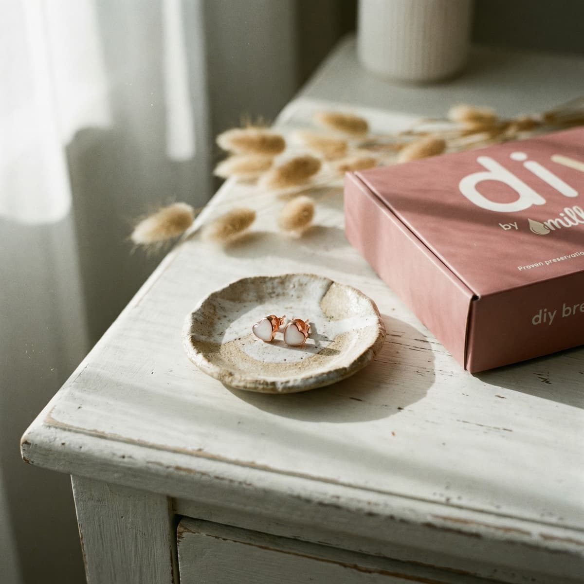 Orecchini a perno con pietra in resina bianca e montatura oro rosa su un piattino accanto alla confezione rosa DIY by MILKIES, kit per creare orecchini latte materno in casa.