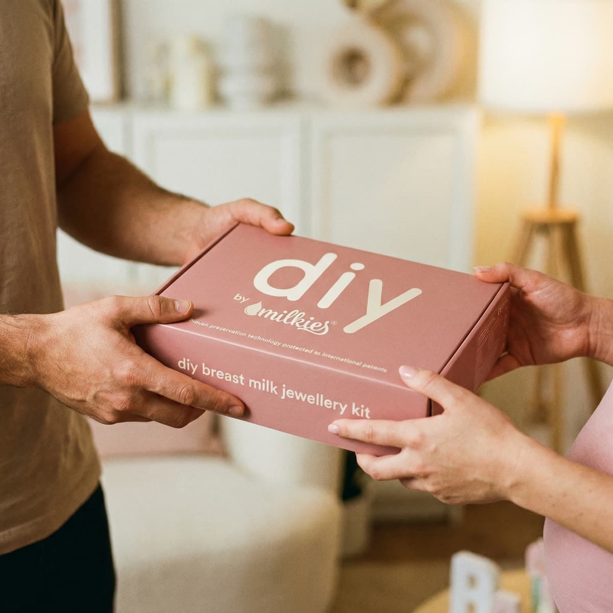 Due mani si scambiano una scatola rosa “DIY by Milkies” con kit per gioielli in latte materno, ideale come regalo allattamento per creare un ricordo personale a casa.