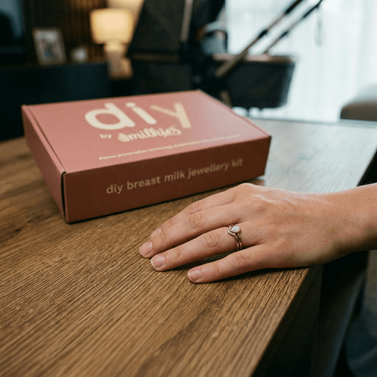 Kit DIY by MILKIES come regalo originale nascita: scatola del kit su tavolo in legno e mano che indossa un anello, per creare gioielli in resina con latte materno.