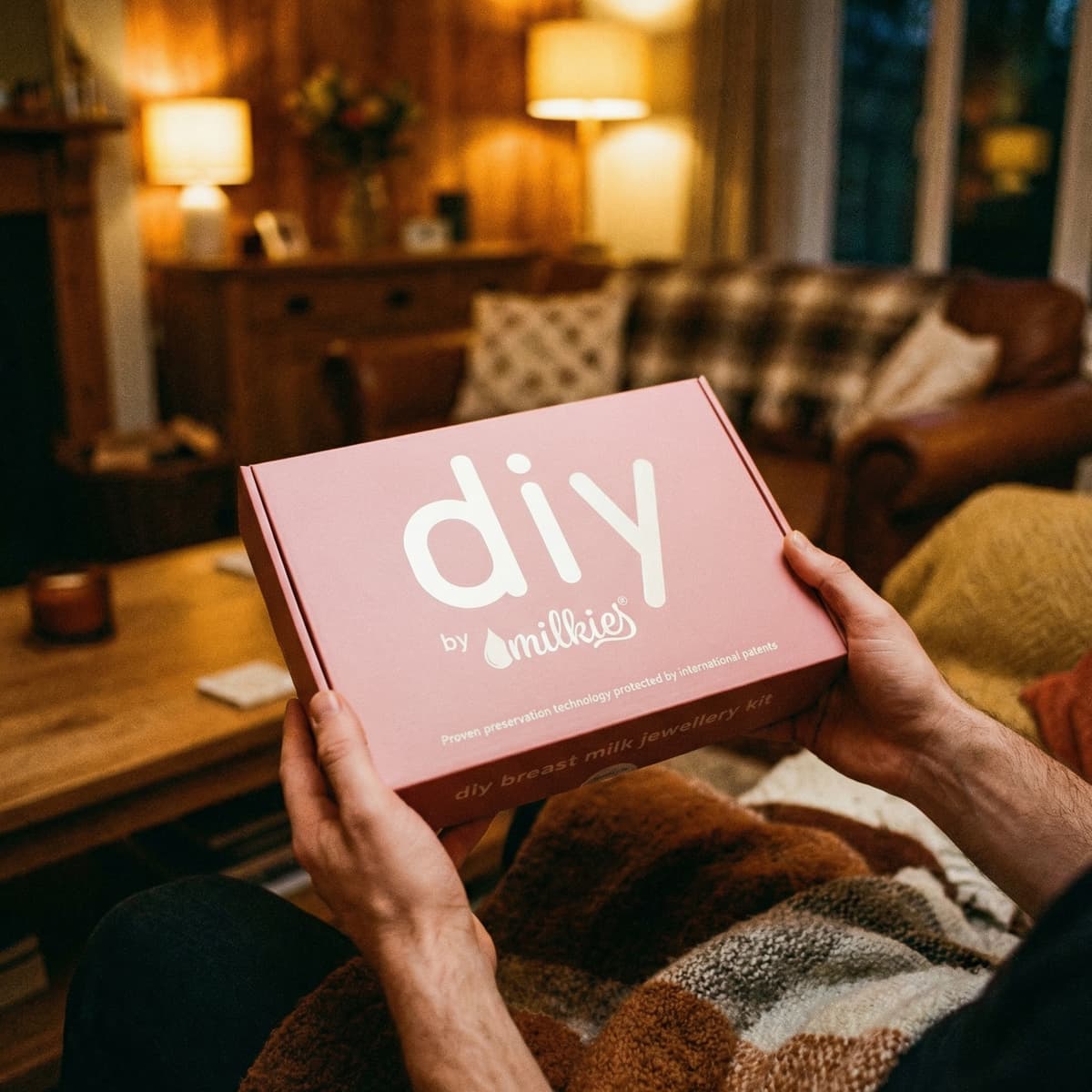 Mani che tengono una scatola rosa “DIY by Milkies” kit fai‑da‑te per creare gioielli con latte materno, idea regalo parto moglie in un salotto accogliente.