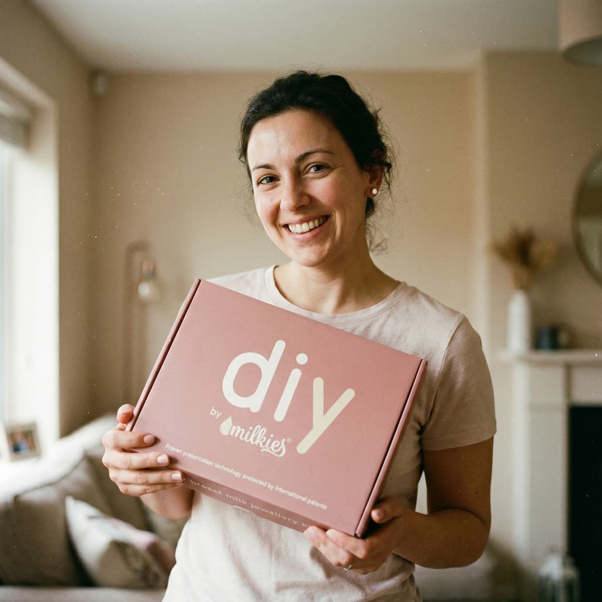 Donna sorridente a casa che mostra la scatola DIY by MILKIES, un kit affidabile e semplice da usare con set per fare orecchini latte materno e altri gioielli ricordo in autonomia.